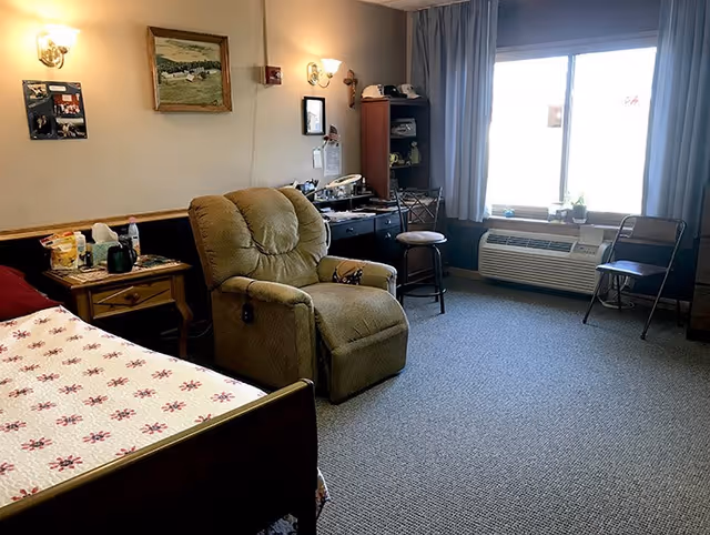 A cozy senior living bedroom featuring a bed with a floral quilt, a beige recliner chair, a wooden nightstand with various items, a desk with a chair and shelves, and a large window with curtains letting in natural light. The room has carpeted flooring and soft wall lighting.