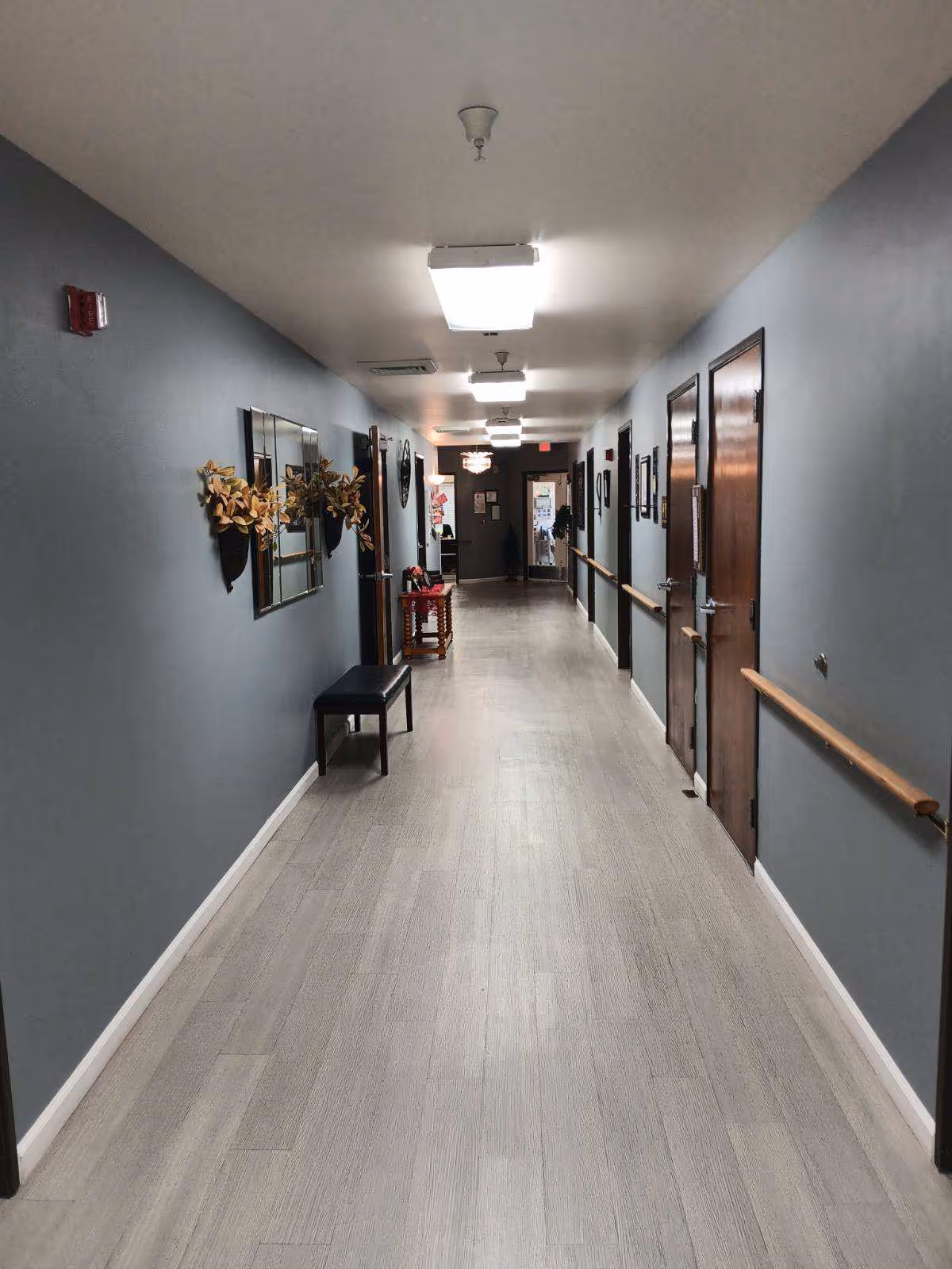 A long, well-lit hallway in a senior living facility with light wood flooring and gray walls. The hallway features wooden handrails on both sides, several closed wooden doors, wall-mounted decorations including mirrors and floral arrangements, and a bench on the left side. The end of the hallway has a door with a window and some visible signage.