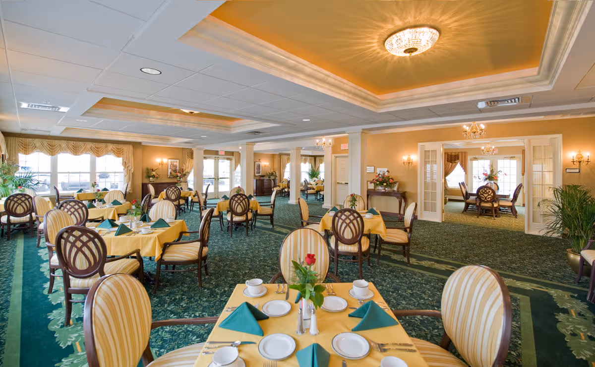 A spacious, well-lit dining room with multiple round tables covered in yellow tablecloths, each set with white plates, cups, and green folded napkins. The room features elegant wooden chairs with striped upholstery, green carpet with floral patterns, large windows with draped curtains, and warm yellow walls. There are chandeliers and wall sconces providing lighting, and some plants and floral arrangements add decoration.