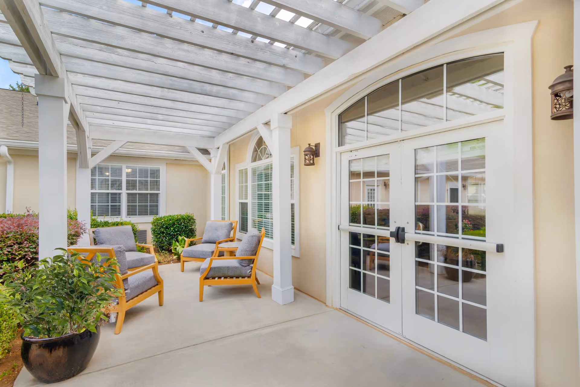 Outdoor patio area with a white pergola overhead, four cushioned wooden chairs arranged around the space, potted plants, and large glass double doors leading inside a building.
