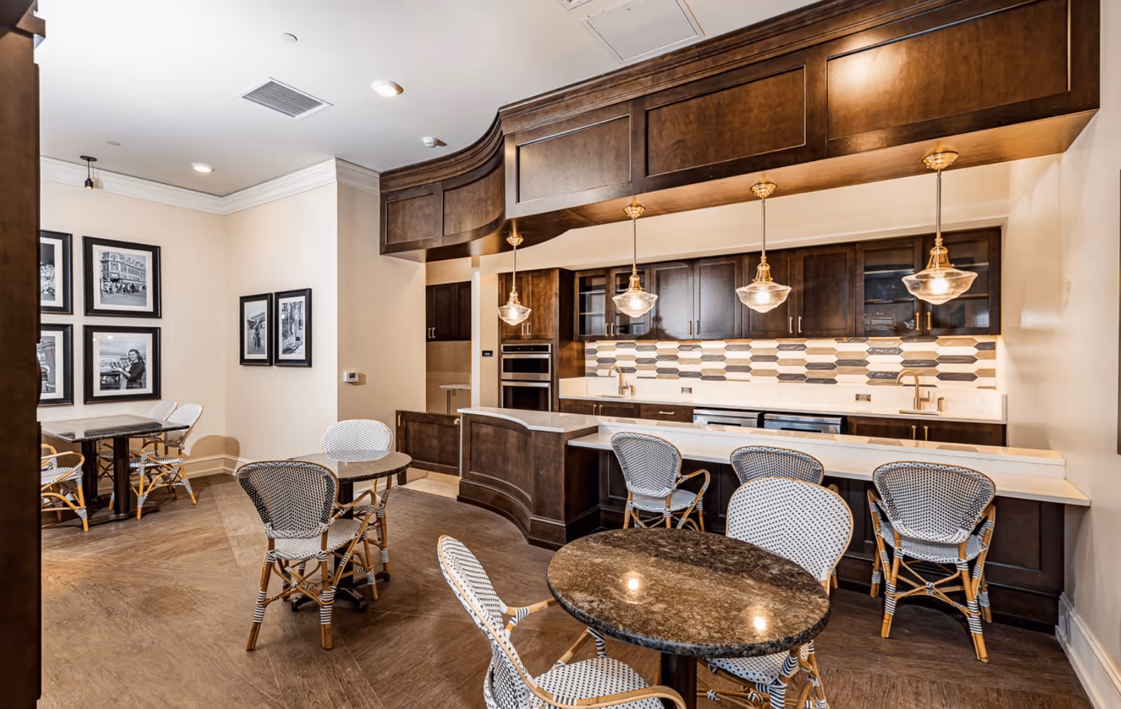 A modern kitchen and dining area with dark wood cabinetry, a white countertop, and a patterned backsplash. There are four pendant lights hanging above the counter and several round tables with wicker chairs arranged in the room. Black and white framed photographs hang on the wall in the background.