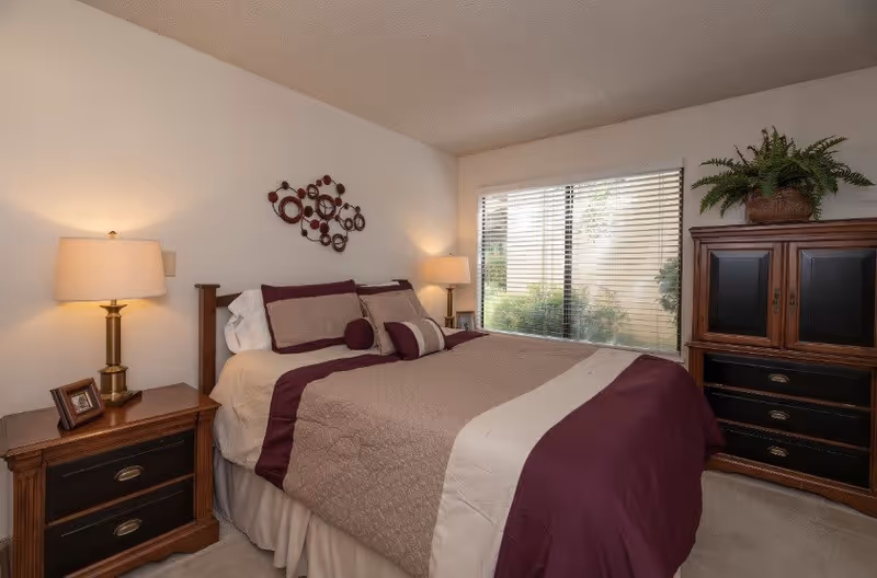 A cozy bedroom with a neatly made bed featuring beige and purple bedding. There are two wooden nightstands on either side of the bed, each with a lamp. A decorative metal wall art hangs above the bed. A large window with blinds lets in natural light, and a wooden dresser with a potted plant on top is positioned near the window.