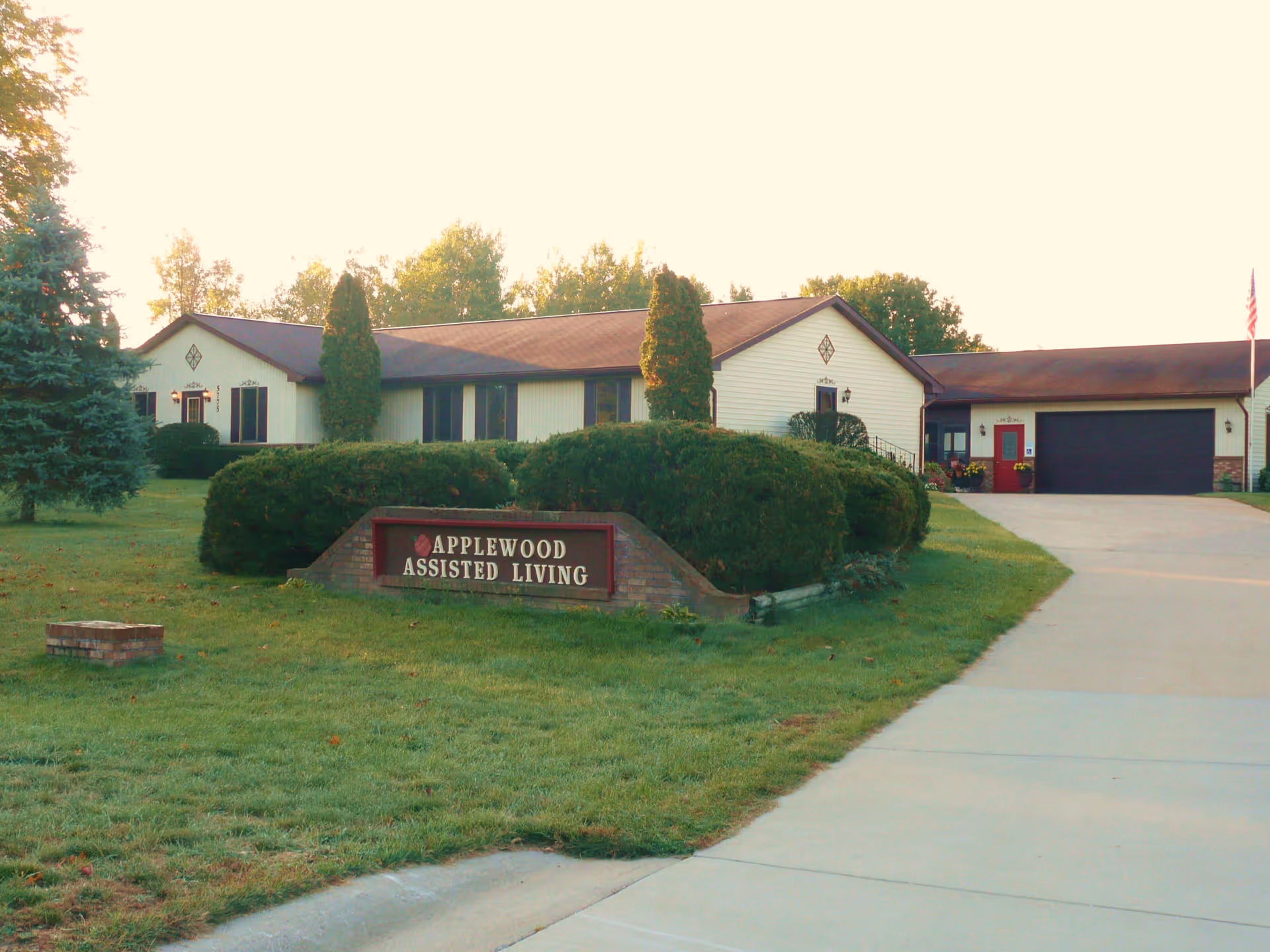Exterior view of Applewood Assisted Living facility with a single-story building, manicured bushes, a lawn, and a driveway leading to a garage. There is a sign in front of the bushes that reads 'Applewood Assisted Living'.