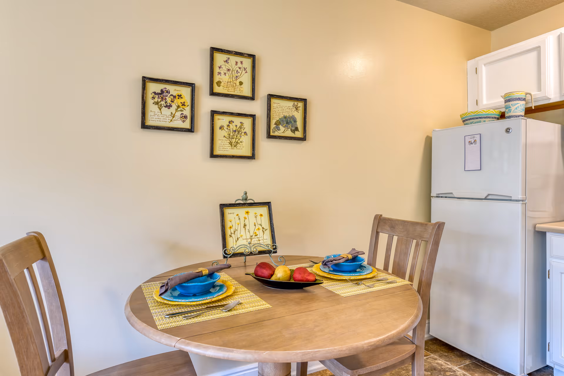 Small dining area with a round wooden table set for two, framed botanical wall art, and a white refrigerator.