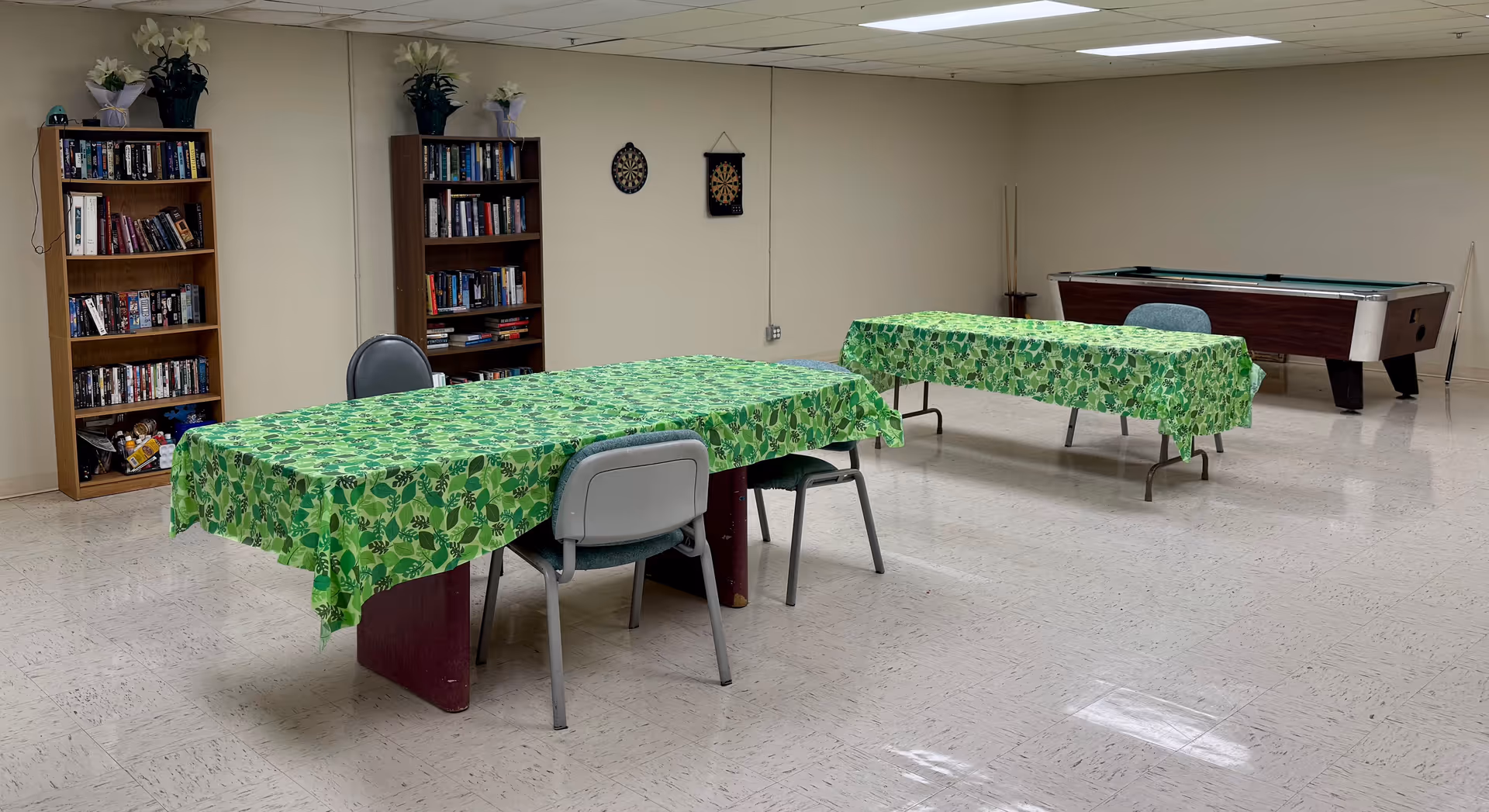 A recreational room with two tables covered in green leaf-patterned tablecloths, each surrounded by chairs. In the background, there are two wooden bookshelves filled with books and decorative plants on top. A dartboard is mounted on the wall, and a pool table is positioned in the corner of the room. The floor is tiled, and the ceiling has fluorescent lighting.