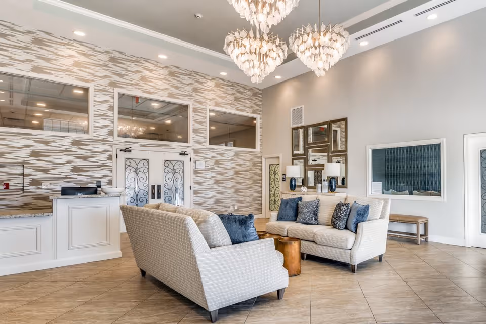 A spacious and elegant senior living facility lobby with two beige sofas arranged around two small wooden tables. The sofas have blue and patterned throw pillows. The walls feature a modern textured design and multiple mirrors. There are two large, ornate chandeliers hanging from the ceiling, and a reception desk is visible on the left side. The floor is tiled with a light brown pattern.