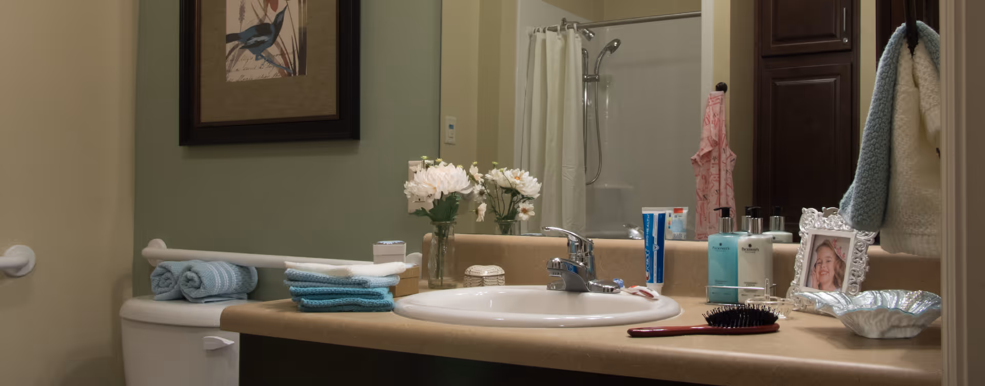 Bathroom sink area with a faucet, a mirror, and various items on the countertop including a hairbrush, soap dispensers, a framed photo of a smiling child, a silver decorative dish, and a vase with white flowers. Towels are folded on the toilet tank, and a shower with a curtain is visible in the background.