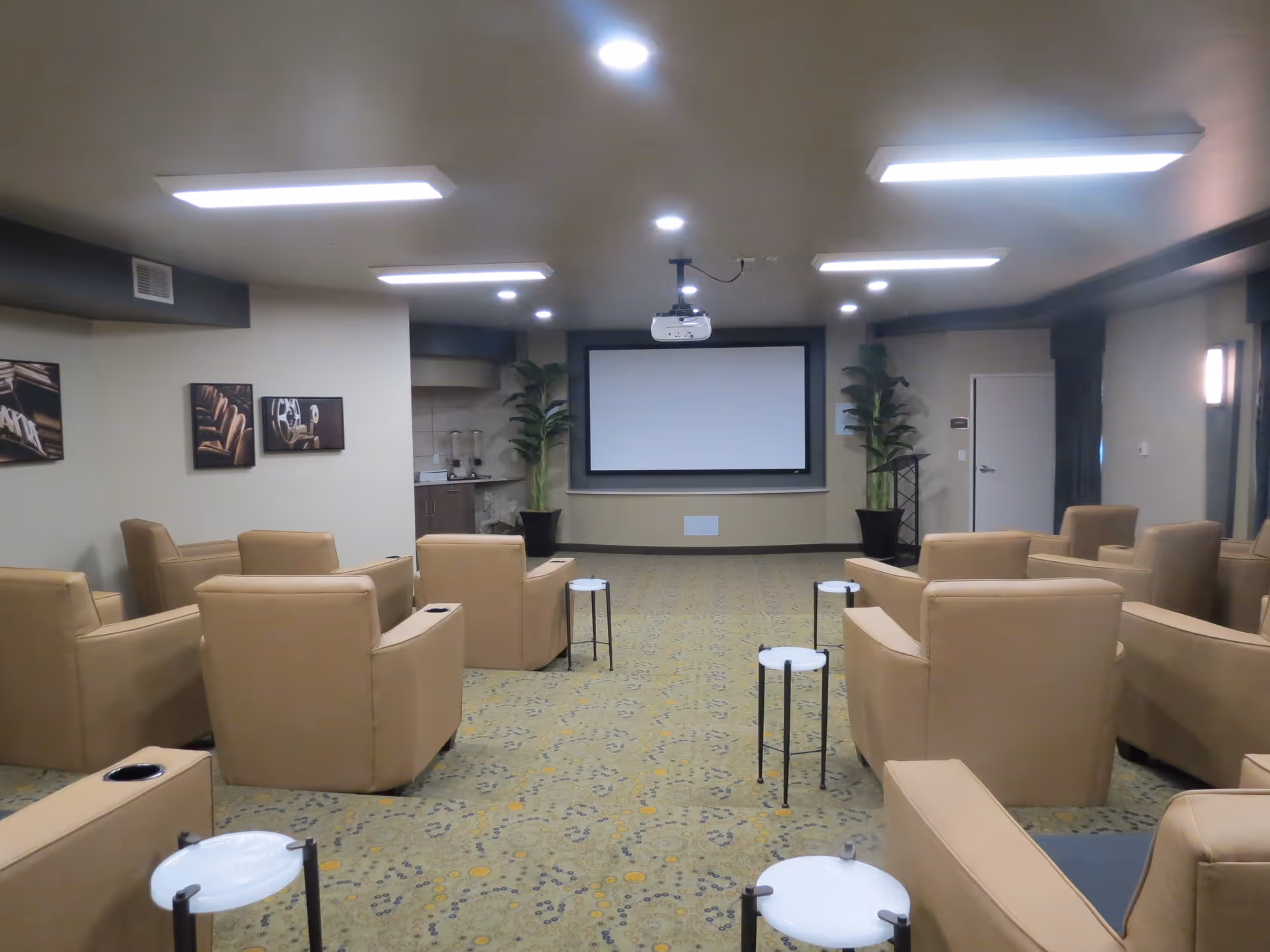 A small theater room with beige recliner chairs arranged in rows facing a large blank projector screen. The room has a patterned carpet, several small round white tables, potted plants on either side of the screen, and framed pictures on the walls. Ceiling lights illuminate the space.