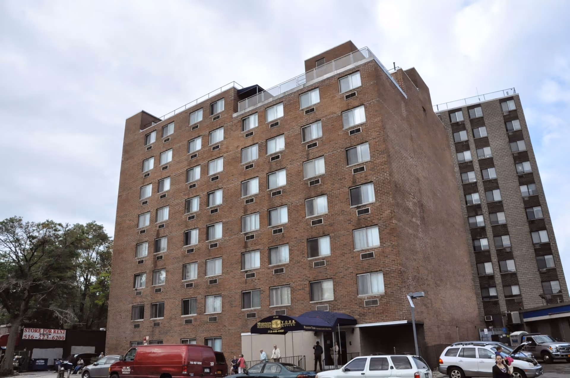 Front view of a multi-story brick apartment/assisted living building with a small entrance canopy and cars parked in front.