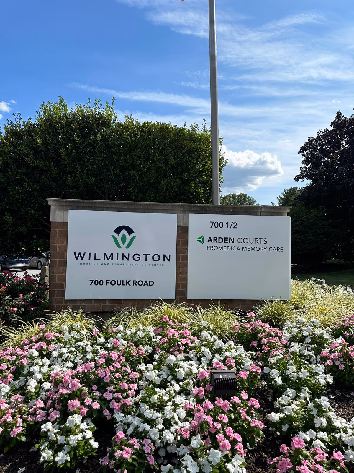 Entrance signage for Wilmington Nursing and Rehabilitation Center and Arden Courts set on a brick sign surrounded by pink and white flowers under a blue sky.