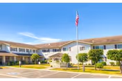 Exterior view of a two-story senior living facility building with a flagpole flying the American flag in front. The building has multiple windows and a covered entrance, surrounded by neatly trimmed bushes and a parking area.