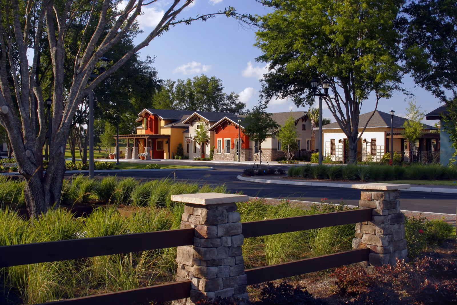 View of a senior living facility named Arlington Ridge showing a landscaped outdoor area with green grass, trees, and a stone and wooden fence in the foreground. In the background, there are several buildings with a mix of red, beige, and stone exteriors under a blue sky with some clouds.