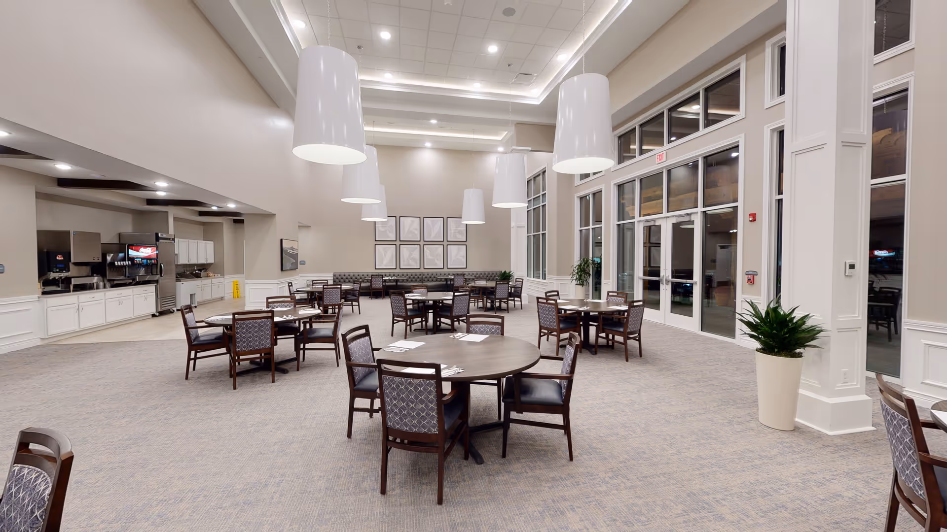 Spacious dining/activity room with round tables and chairs, high ceilings and pendant lights, a buffet counter to the left, and large windows on the right.