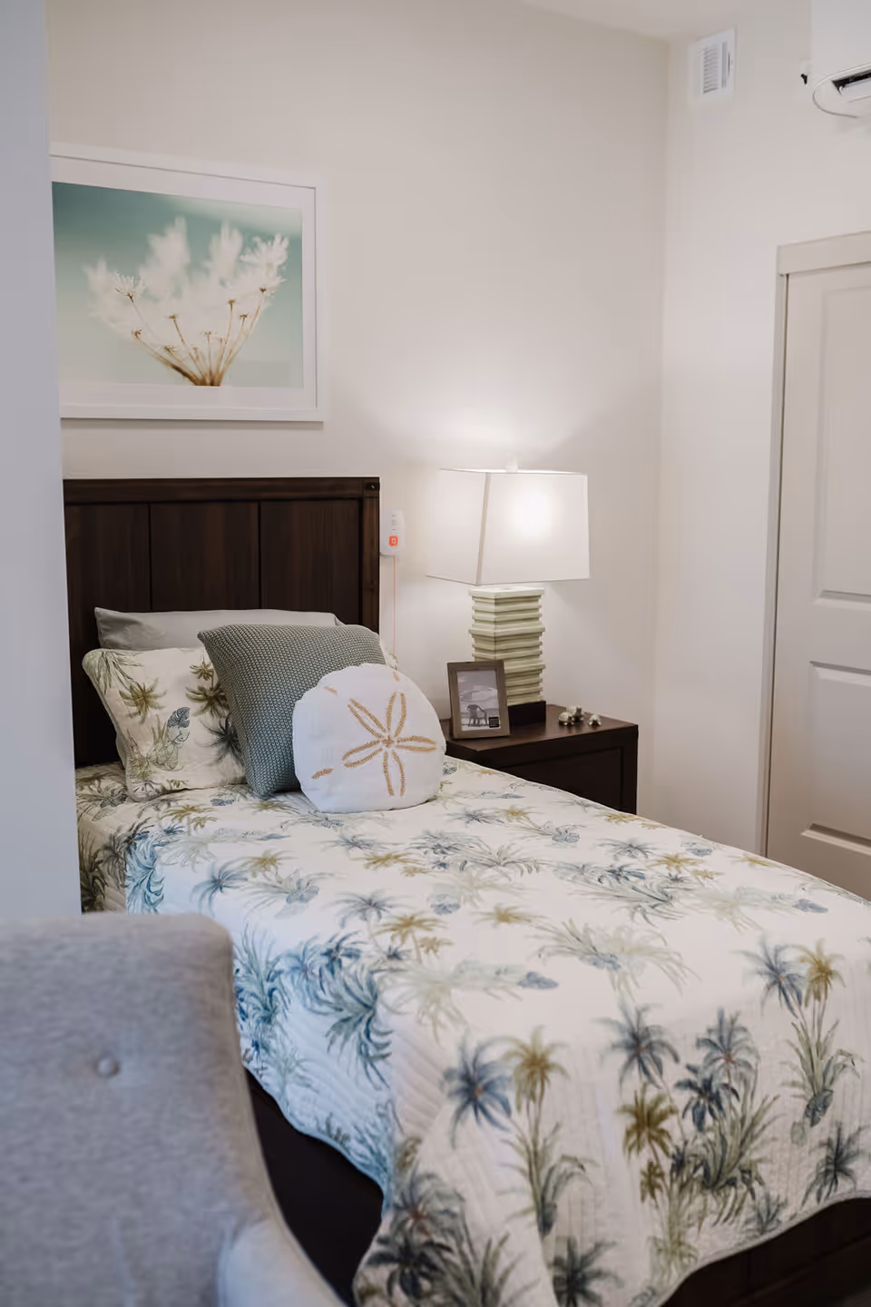 Single bedroom with a twin bed dressed in botanical bedding, a wooden headboard, and a bedside table with a lamp and framed photo.