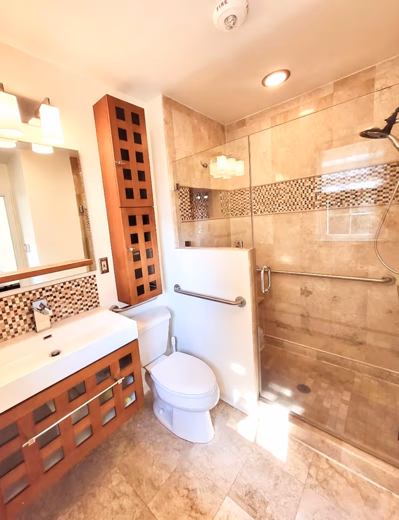 A modern bathroom featuring a white toilet, a rectangular sink with a wooden cabinet below, and a tall wooden storage cabinet mounted on the wall. The shower area has a glass door and beige tiled walls with a decorative mosaic tile strip. There are grab bars installed near the toilet and inside the shower for safety. The bathroom is well-lit with ceiling lights and a wall-mounted light fixture above the mirror.
