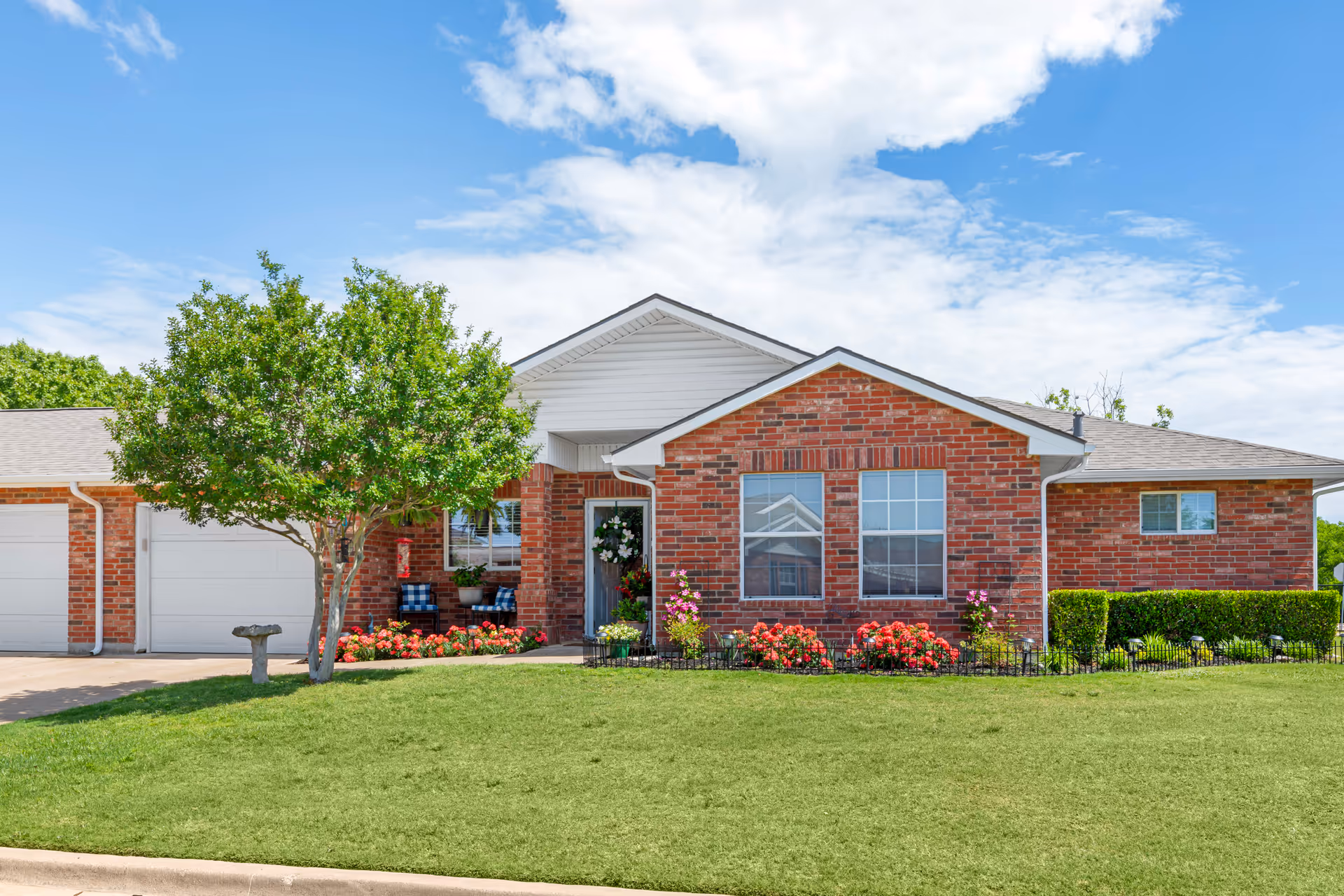 Front exterior view of a single-story brick building with a well-maintained lawn, a tree, flower beds with red and pink flowers, a birdbath, and a small porch with two chairs and potted plants under a partly cloudy blue sky.