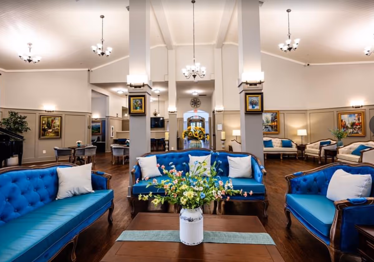 Spacious, elegant communal seating area with blue tufted sofas around a coffee table topped with a floral arrangement, chandeliers, and additional seating in the background.