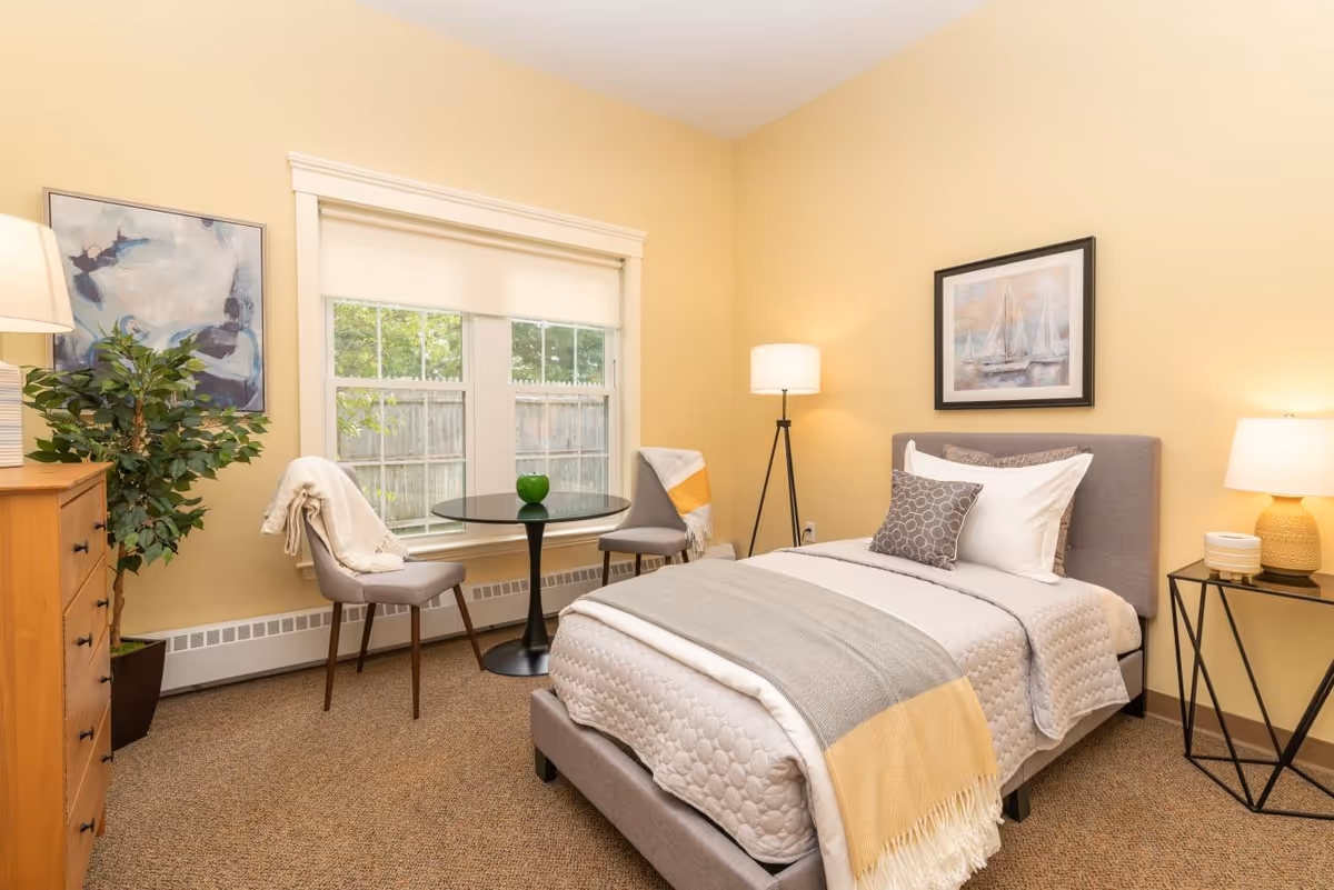 A cozy bedroom with a single bed dressed in white and gray bedding with a yellow throw blanket. There is a small round black table with two gray chairs, each draped with a blanket, positioned near a window with white blinds. A wooden dresser with a lamp and a potted plant is on the left side, and a floor lamp and a side table with a lamp are on the right side. The walls are painted light yellow and decorated with framed artwork.