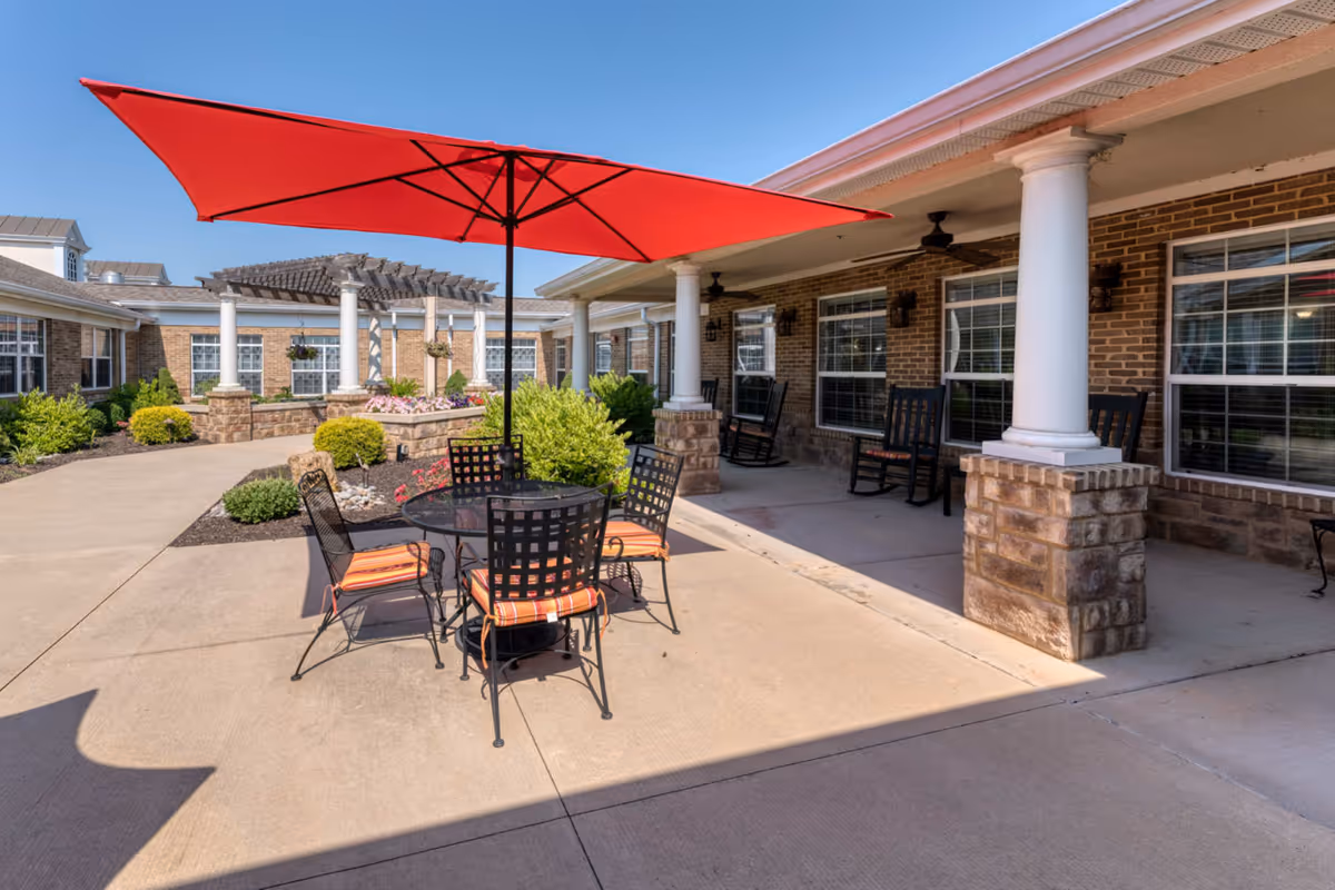 Outdoor patio area at Addington Place of Shoal Creek featuring a round metal table with four chairs that have orange striped cushions, shaded by a large red umbrella. The patio is surrounded by a brick building with white columns and rocking chairs along the covered walkway. There are landscaped bushes and flowers in the background under a clear blue sky.