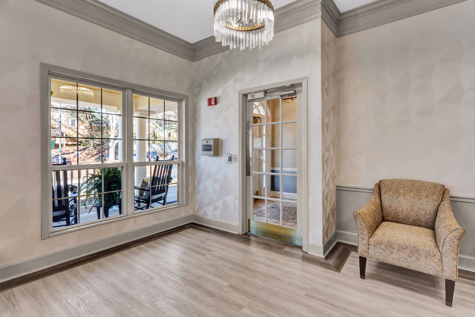 A bright interior corner of a facility with light-colored patterned walls, a large window showing an outdoor porch with rocking chairs and plants, a glass-paneled door, a beige patterned armchair, and a modern chandelier hanging from the ceiling.