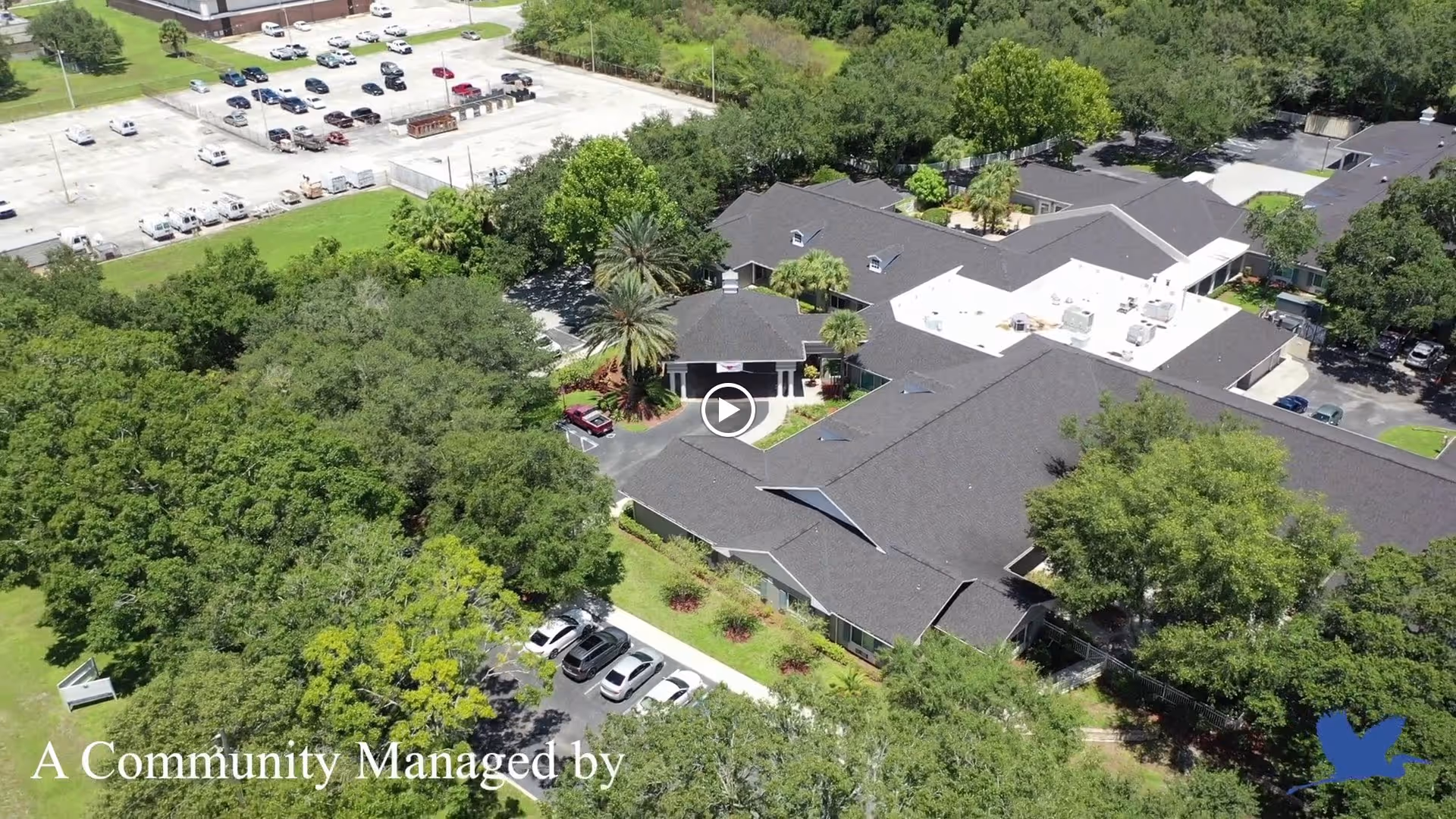 Aerial view of The Colonnade at Northdale senior living facility surrounded by trees and greenery, with parking lots visible nearby and multiple connected buildings with dark roofs.