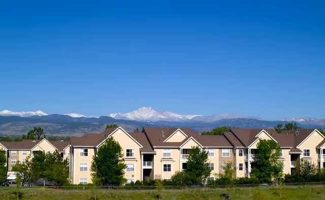 Exterior view of a multi-story residential building with beige siding and brown roofs, surrounded by green trees and grass, with snow-capped mountains visible in the background under a clear blue sky.