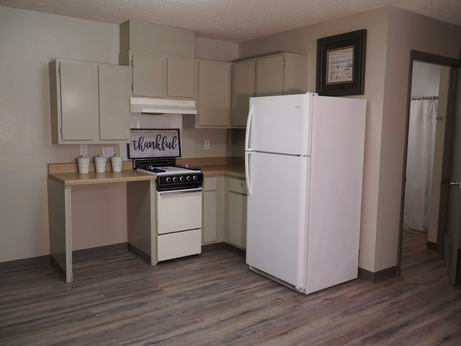 A small kitchen area with beige cabinets, a white refrigerator, a white stove with an oven, and a countertop with three white containers. There is a framed sign on the countertop that says 'thankful'. The floor is wood-patterned, and there is a doorway leading to another room with a shower curtain visible.