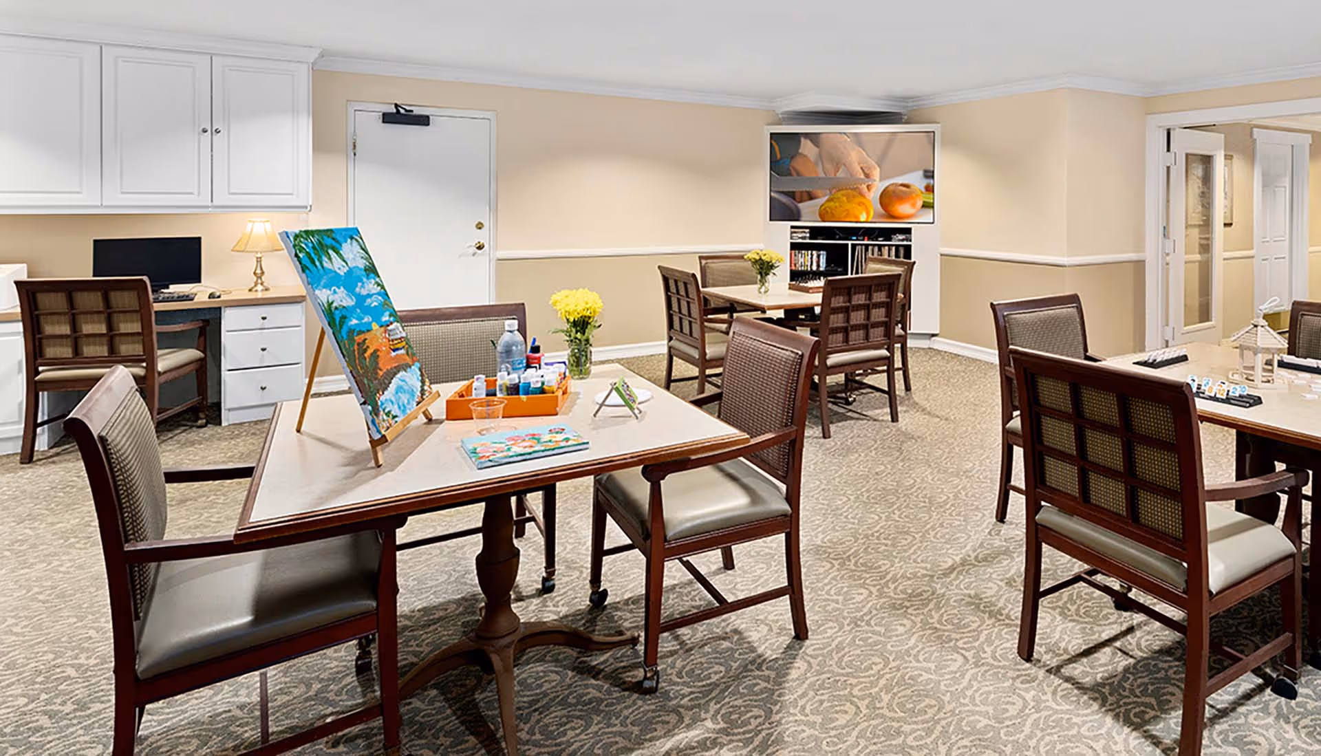 A bright interior activity room with tables and chairs, an easel and art supplies, a TV, and a desk in a senior living facility.