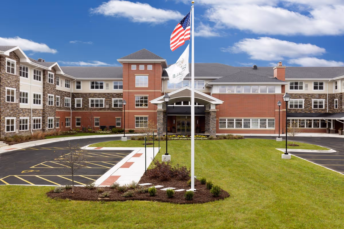 Front exterior view of Clarendale of Algonquin, a multi-story senior living facility with a combination of brick and stone facade, multiple windows, a covered entrance, an American flag and another flag on flagpoles, surrounded by a well-maintained lawn and parking spaces.