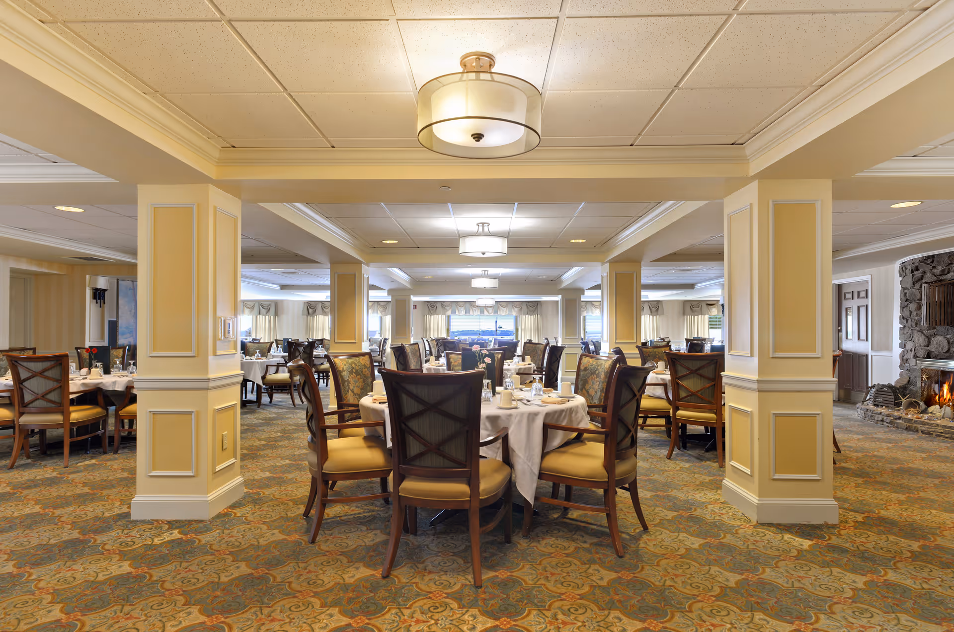 A spacious dining room with multiple round tables covered with white tablecloths and set with cups, plates, and silverware. The room features patterned carpet, cream-colored walls with decorative molding, and several columns. There are cushioned wooden chairs around the tables and a stone fireplace with a fire burning on the right side of the room. Large windows with curtains allow natural light to enter the space.