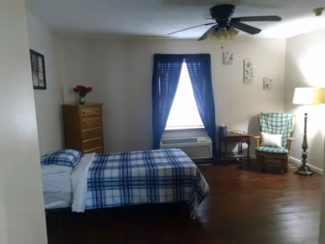 A bedroom with a single bed covered in blue and white plaid bedding, a wooden dresser with a vase of red flowers, a window with dark blue curtains, an air conditioning unit below the window, a wooden rocking chair with a green plaid cushion, a small side table, and a floor lamp. The room has wooden flooring and a ceiling fan with lights.