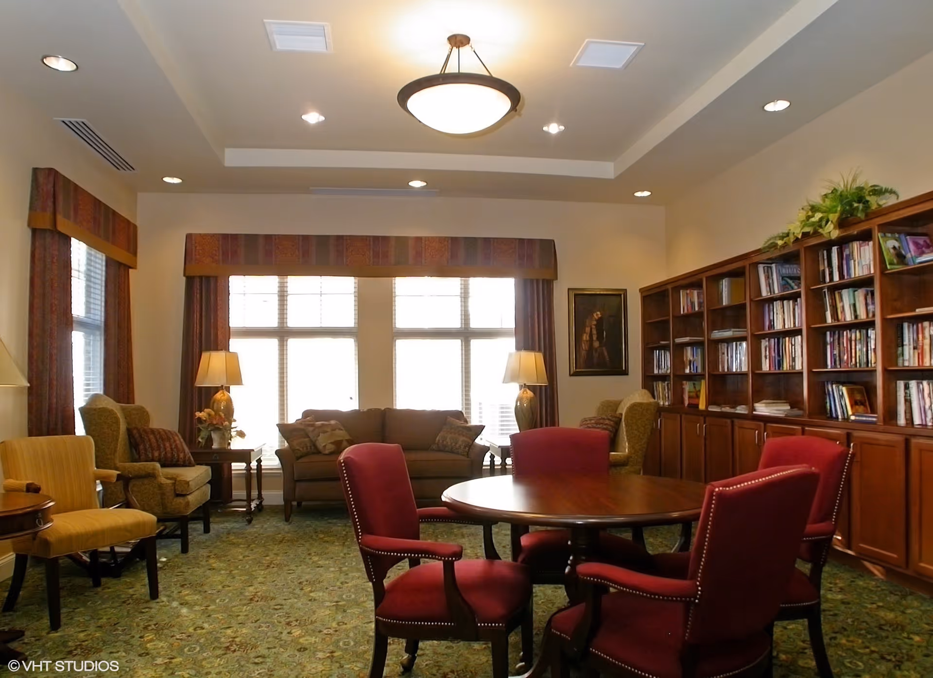 A cozy living room area with a round wooden table surrounded by four red upholstered chairs in the foreground. In the background, there is a brown sofa flanked by two side tables with lamps, two armchairs, and a large wooden bookshelf filled with books and decorative plants. The room has large windows with valances and curtains, recessed ceiling lights, and a central hanging light fixture.