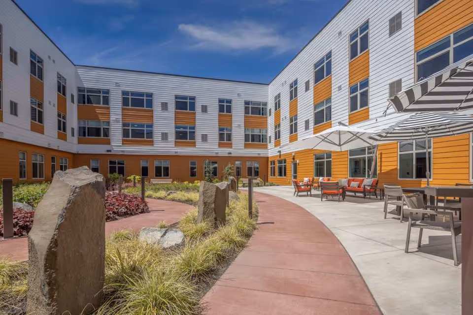 Outdoor courtyard area of a senior living facility with a curved red walking path, landscaped plants, large standing stones, and seating areas with tables, chairs, and umbrellas. The building surrounding the courtyard has white and orange paneling with many windows under a clear blue sky.