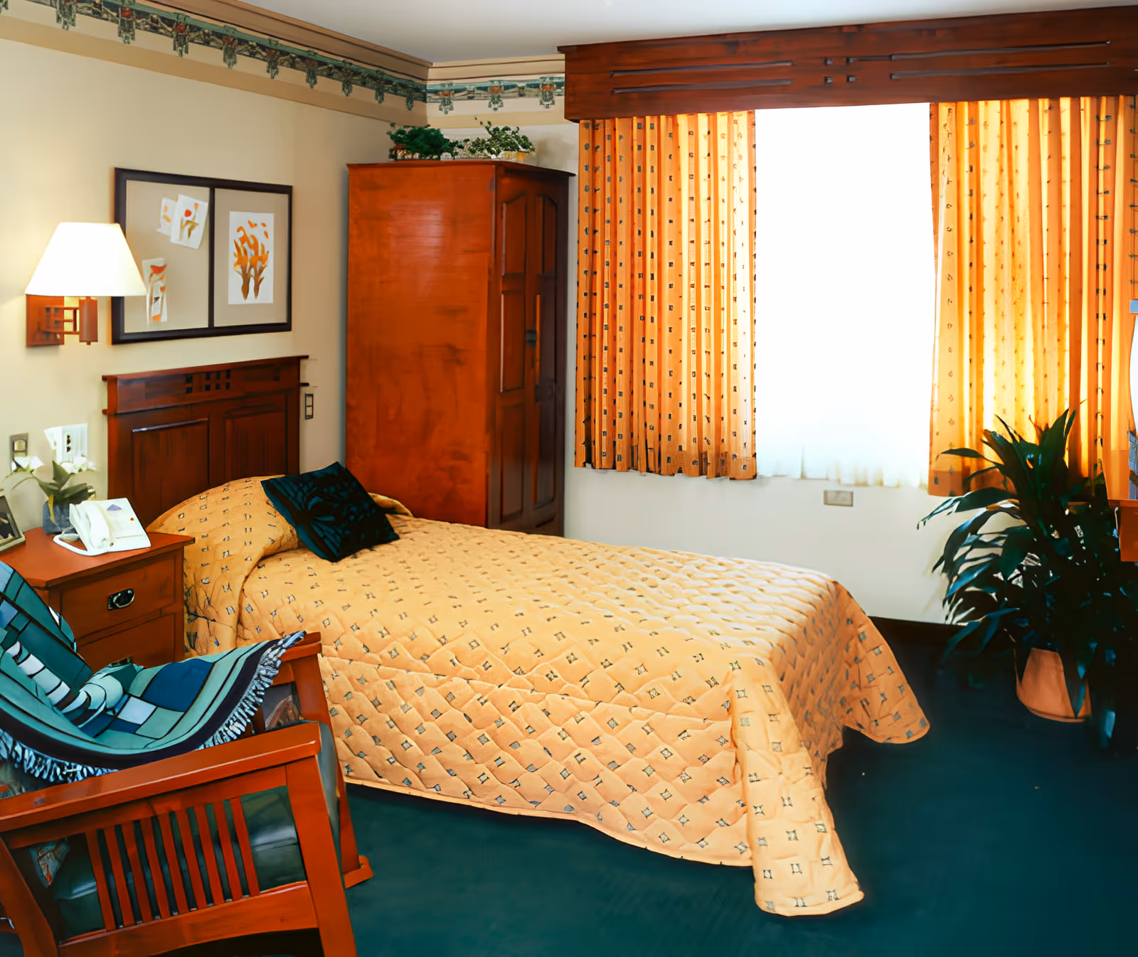 A cozy bedroom with a single bed covered in a yellow quilted bedspread, a wooden headboard, and a dark green pillow. Next to the bed is a wooden nightstand with a lamp and a telephone. There is a wooden armchair with a green and blue striped blanket draped over it. A large wooden wardrobe stands against the wall, and a window with yellow patterned curtains lets in bright light. A green potted plant is placed near the window, and framed artwork hangs on the wall above the bed.