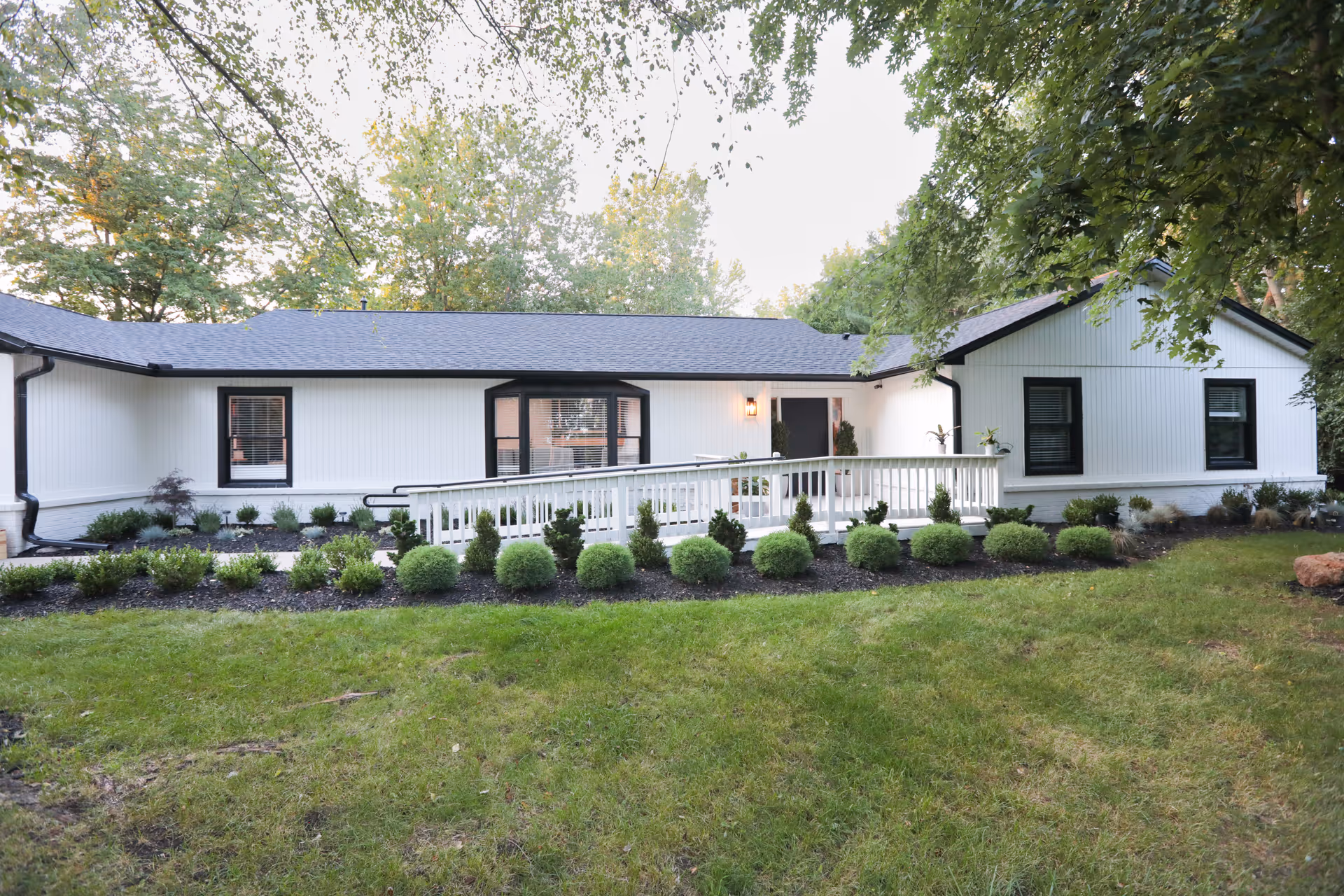 Exterior view of a single-story white building with black trim, surrounded by green grass and landscaped bushes. There is a white ramp leading to the entrance, and trees are visible in the background.
