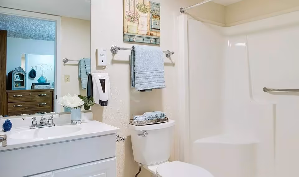 A clean and well-lit bathroom featuring a white sink with a silver faucet, a large mirror, a white toilet with a tray holding folded towels and a box of tissues on top, a towel rack with a blue towel, a wall-mounted soap dispenser, and a white shower area with a built-in seat and grab bar.