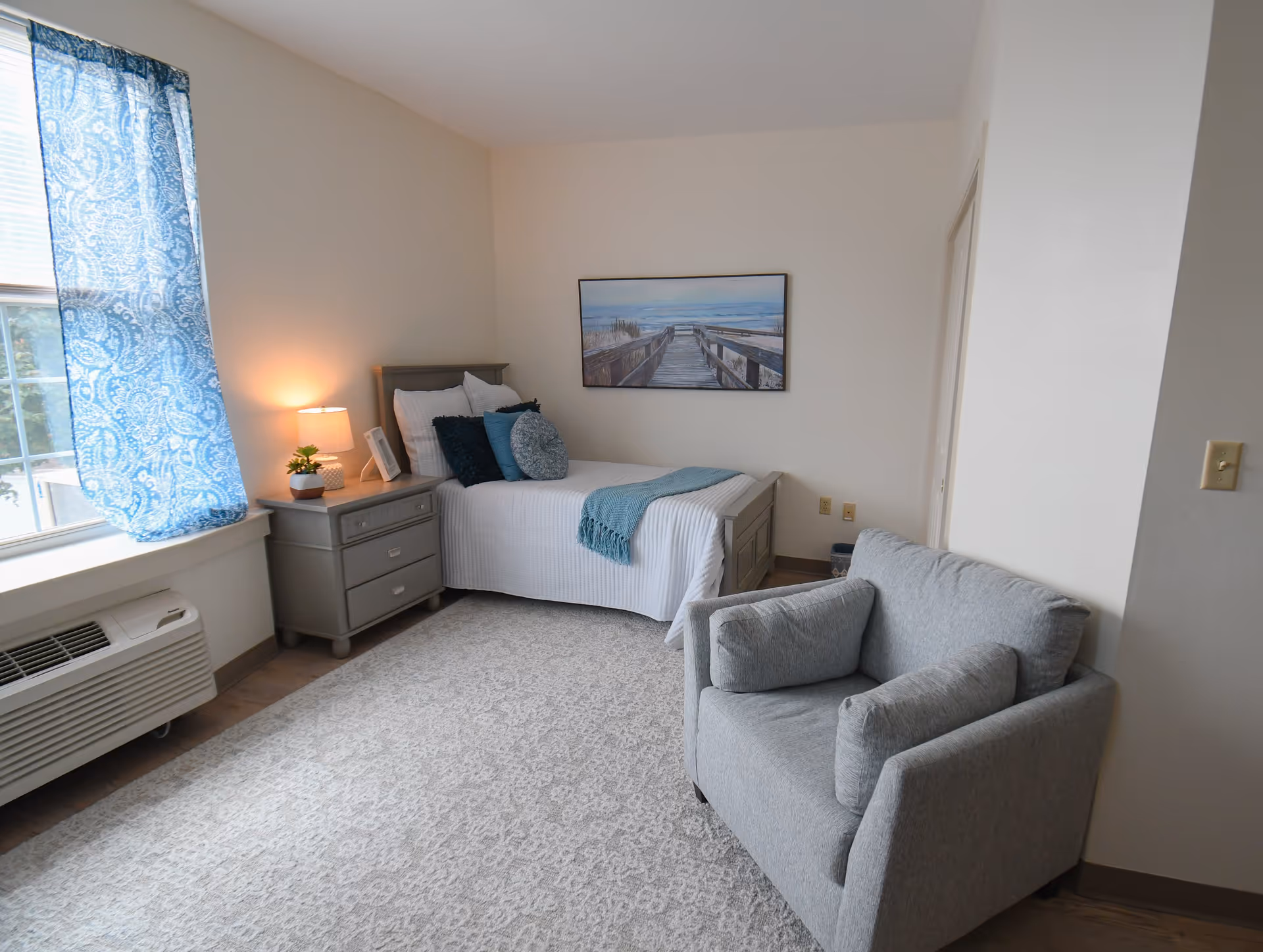 A cozy bedroom with a single bed adorned with white bedding and blue accent pillows and throw. Next to the bed is a gray nightstand with a lamp, a small plant, and a picture frame. A large window with blue patterned curtains lets in natural light. A comfortable gray armchair with cushions is positioned near the bed. The walls are light-colored, and a framed picture of a boardwalk leading to the beach hangs above the bed. The floor is covered with a light patterned rug.
