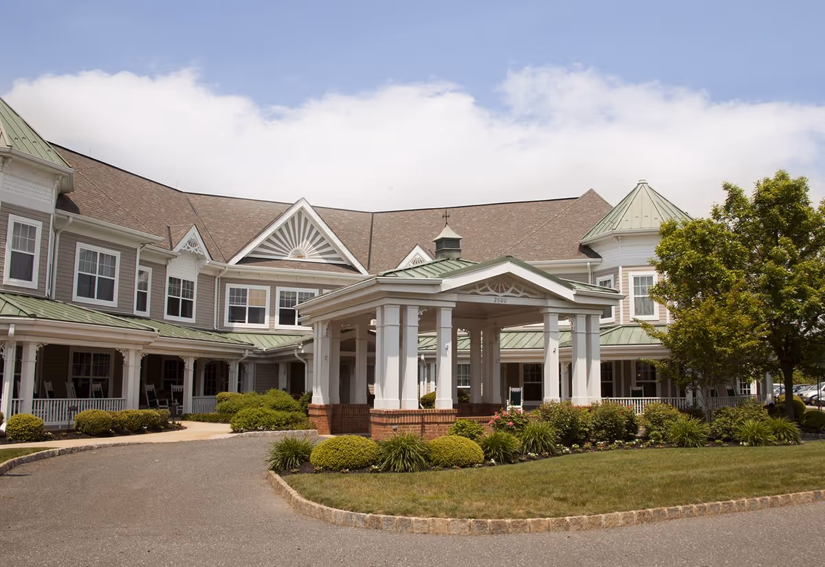 Exterior view of a senior living facility with a large covered entrance, manicured bushes, and a green lawn under a partly cloudy sky.