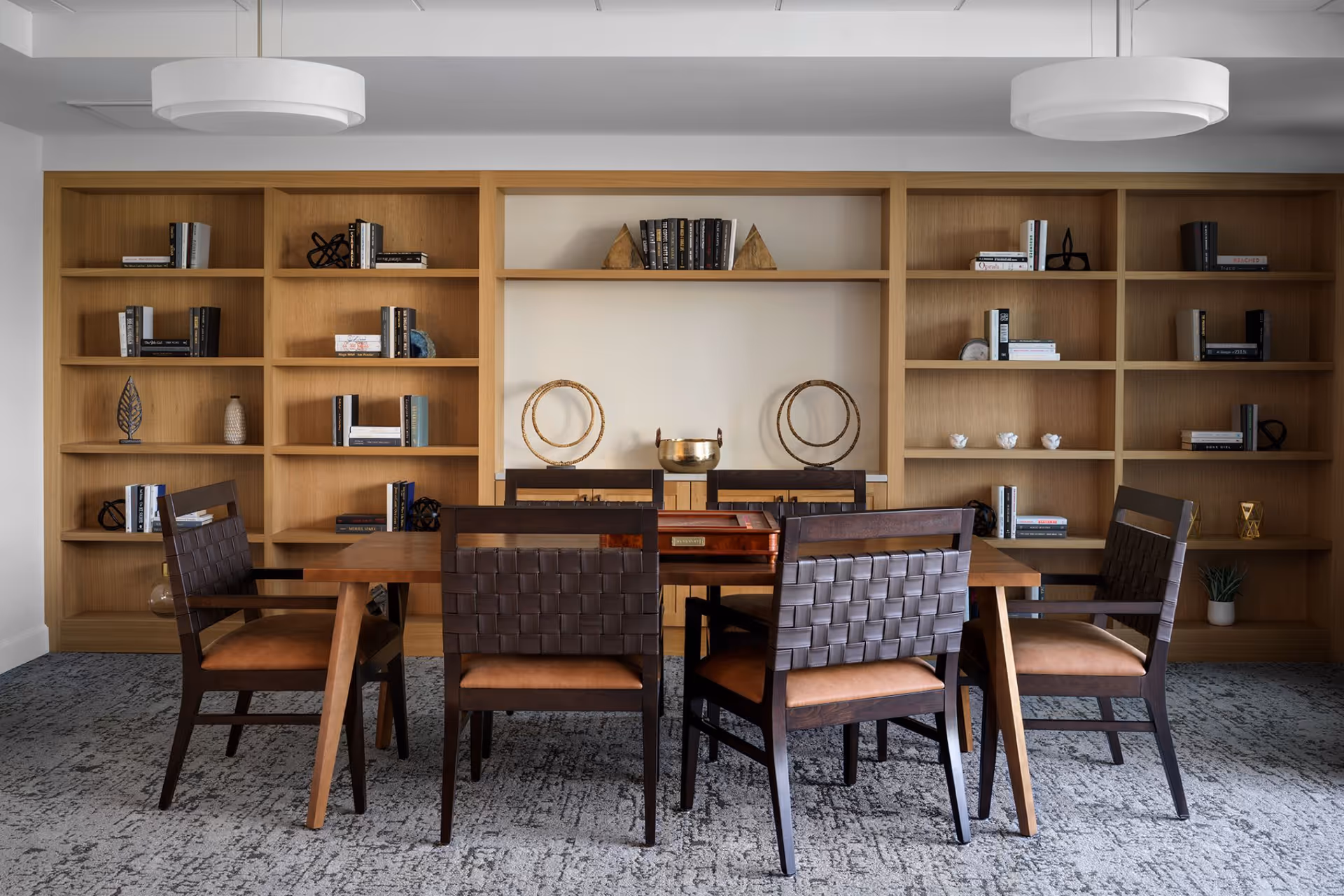 A modern room with a wooden table surrounded by six dark brown chairs with woven backs and tan cushions. Behind the table is a large built-in wooden bookshelf filled with books and decorative items. The room has a gray patterned carpet and two circular white ceiling lights.