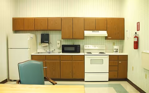 A small kitchen area with wooden cabinets, a white refrigerator, a microwave, a white stove with an oven, a coffee maker, a sink, and a green chair in front of the counter.