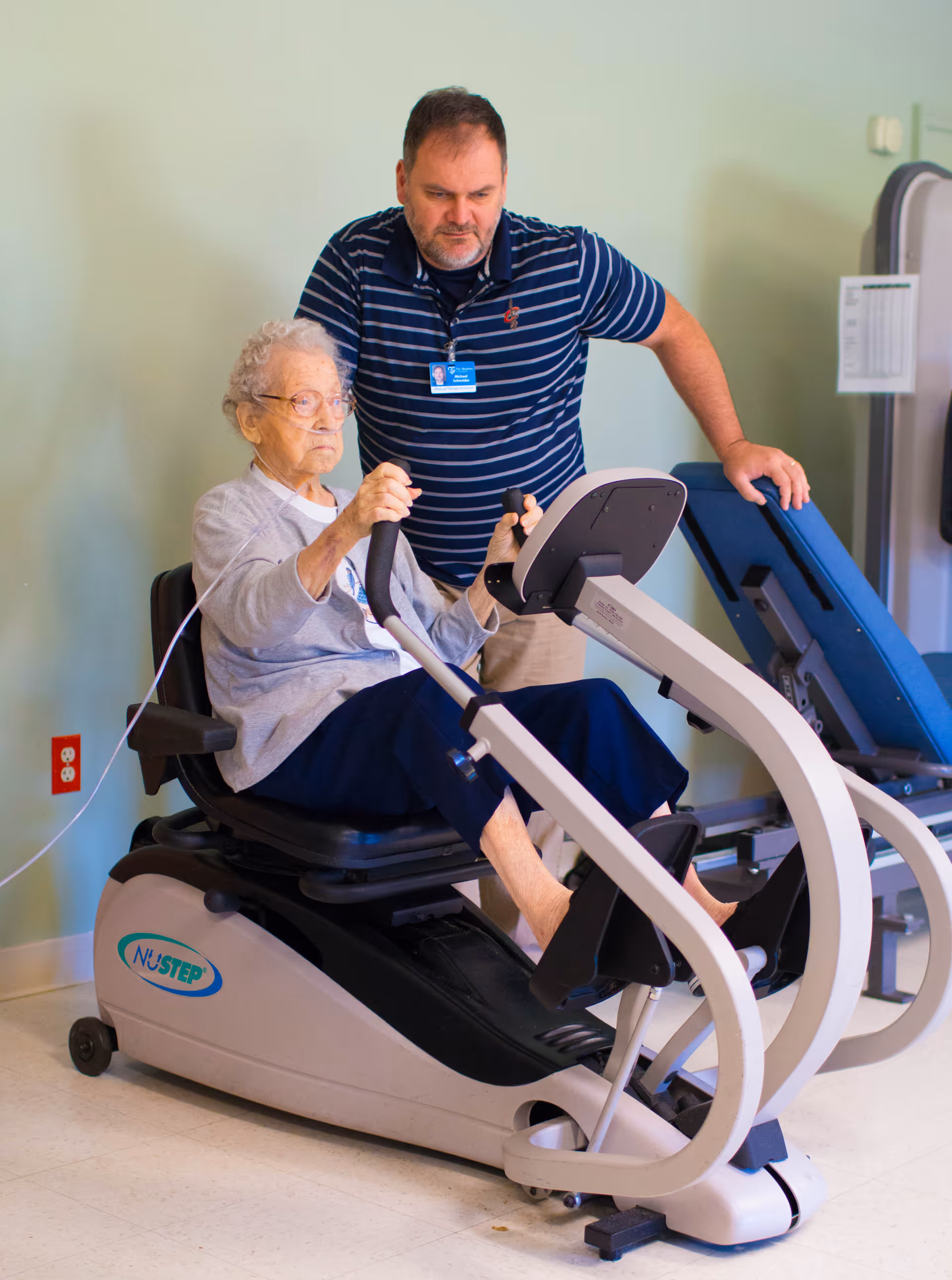 An elderly woman is seated on a NuStep exercise machine, using the handles and pedals for a workout. A man wearing a striped polo shirt and a name badge is standing beside her, providing assistance or supervision. The setting appears to be an indoor fitness or rehabilitation area with exercise equipment in the background.