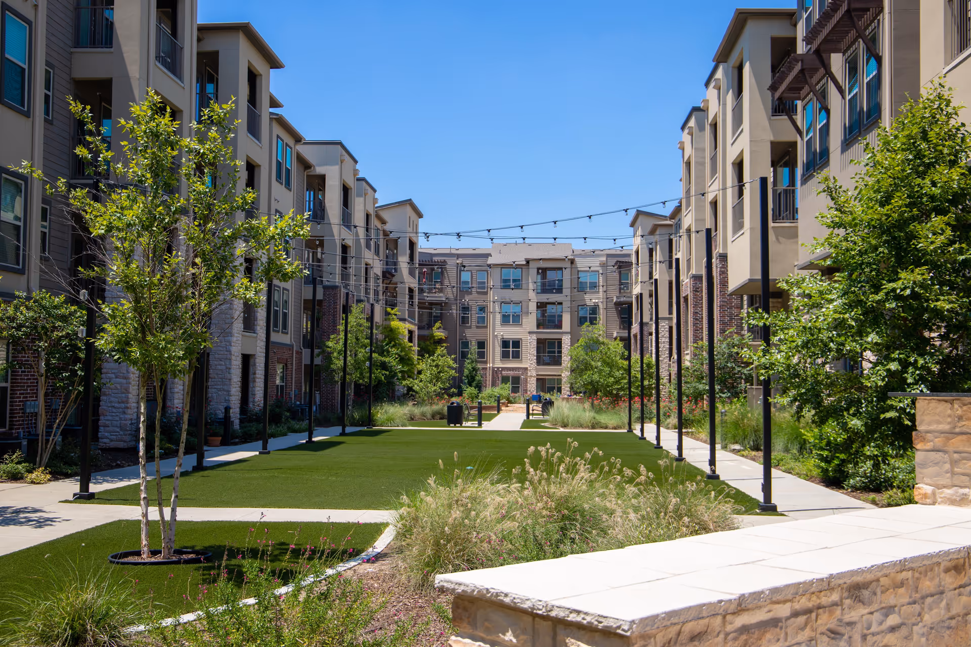 Outdoor courtyard area of a senior living facility with green lawns, young trees, shrubs, and string lights hanging overhead between multi-story beige and brown buildings under a clear blue sky.
