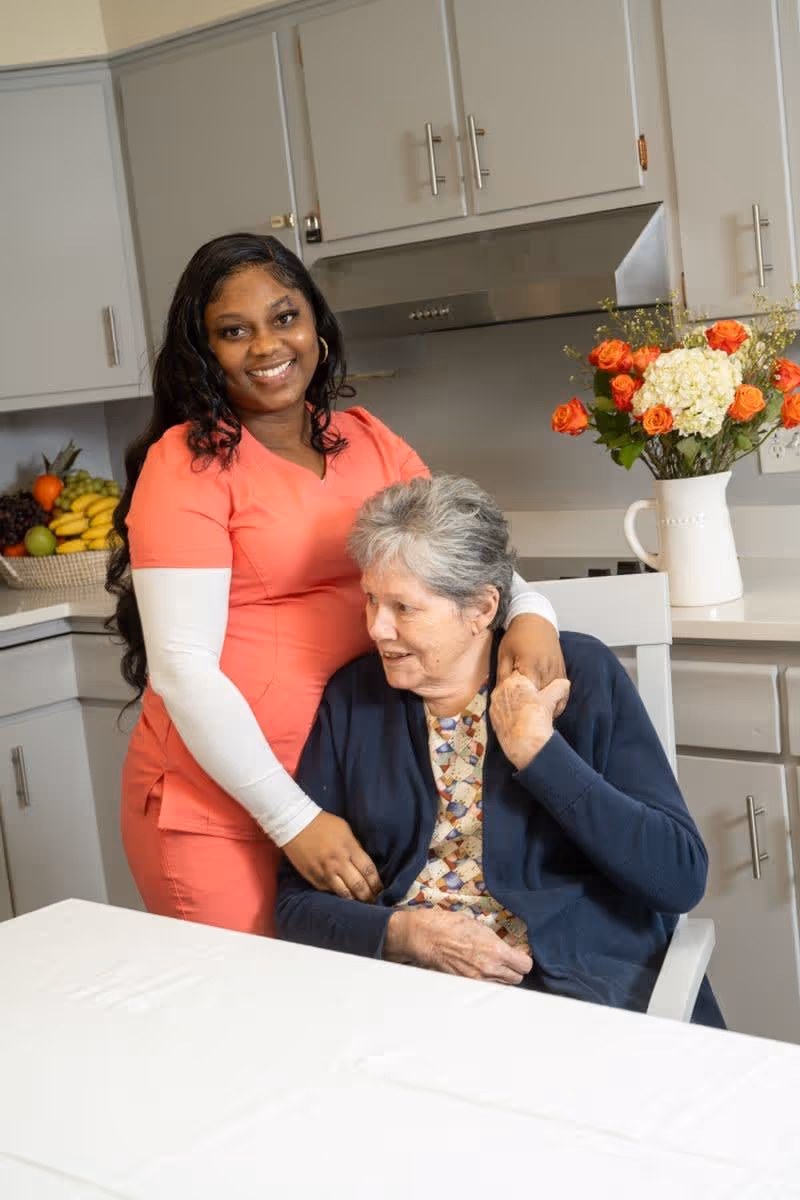 A smiling caregiver in a coral scrub top stands behind an elderly woman seated at a kitchen table. The caregiver has her arm around the elderly woman, who is wearing a patterned blouse and navy cardigan. The background shows a kitchen with white cabinets, a basket of fruit, and a vase of orange and white flowers on the counter.