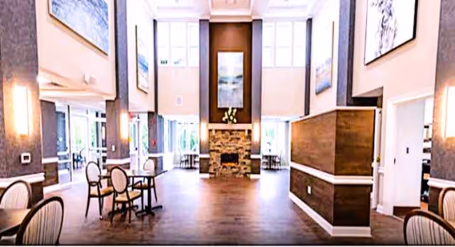 A spacious and well-lit common area with high ceilings, featuring a stone fireplace centered on the far wall. The room has several round tables with chairs arranged around them, large windows allowing natural light, and artwork hanging on the walls. The flooring is wooden, and the overall decor is warm and inviting.