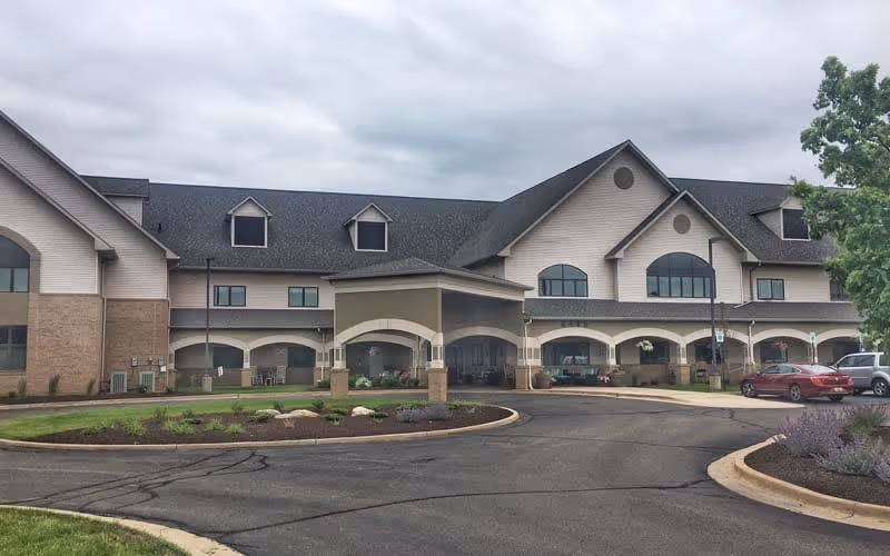 Exterior front view of a two-story retirement center building with a covered entrance, multiple windows, and a circular driveway with parked cars and landscaped areas under a cloudy sky.
