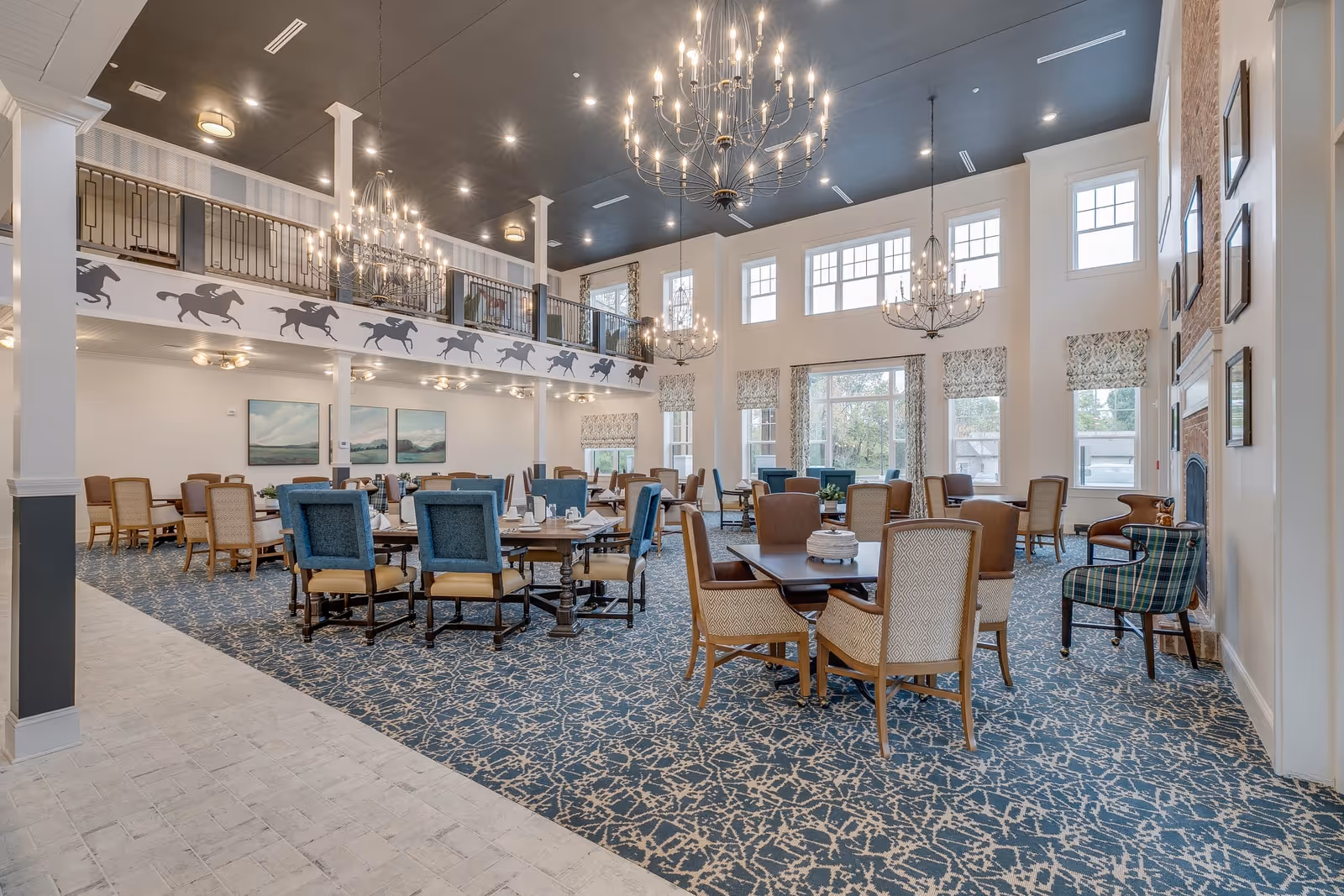 Spacious and elegant dining room with multiple tables and chairs arranged on a patterned blue carpet. The room features high ceilings with large windows allowing natural light, several chandeliers, and a balcony with horse silhouette decorations. The walls are adorned with framed artwork and a brick fireplace is visible on the right side.