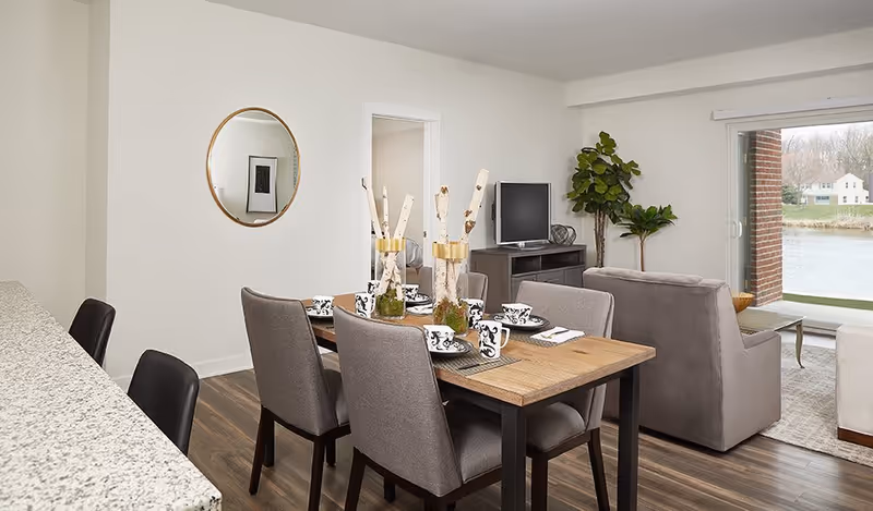 A modern senior living dining and living area with a wooden dining table set for six, gray upholstered chairs, a granite countertop with black bar stools, a gray sofa, a TV on a stand, a large potted plant, a round wall mirror, and a sliding glass door overlooking a water view outside.