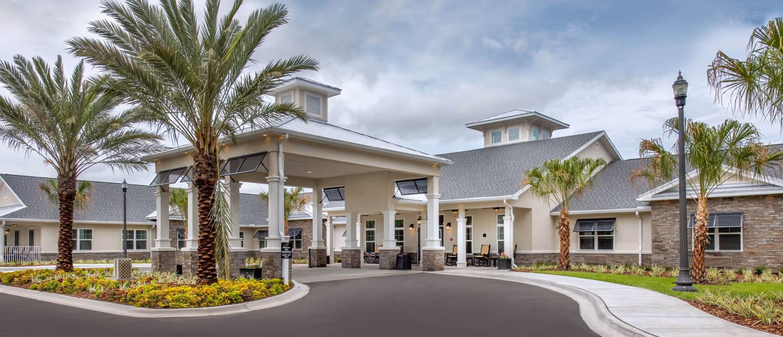Exterior view of The Addison of Oviedo senior living facility showing a covered entrance with columns, palm trees, landscaped garden beds, and a paved driveway under a cloudy sky.