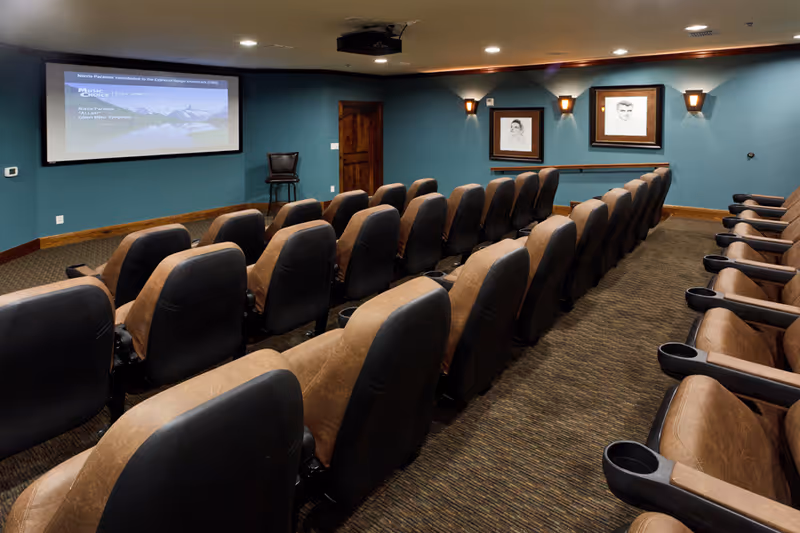 A small theater room with multiple rows of brown and black cushioned seats facing a large projection screen on a teal-colored wall. The room has carpeted flooring, two framed black and white portraits on the right wall, and wall-mounted lights above the portraits. A wooden door and a single chair are visible near the screen.