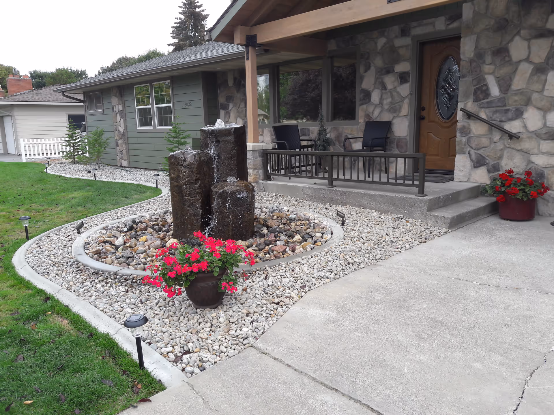 Front entrance of a stone-faced retirement home with a three-column water fountain, rock landscaping, and potted red flowers.